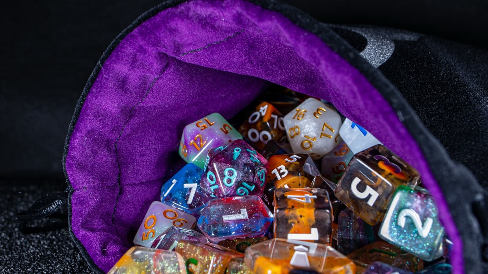 A black velvet dice bag with a purple lining, spilling over with colorful resin dice